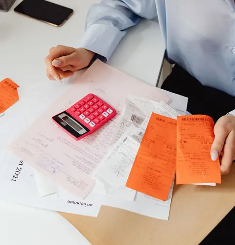 Hands sorting orange receipts beside a pink calculator and a stack of invoices