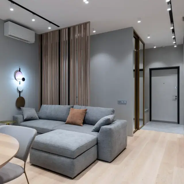 A bright condo living room with a grey sofa, a round dining table and a white wall-mounted aircon above the doorway