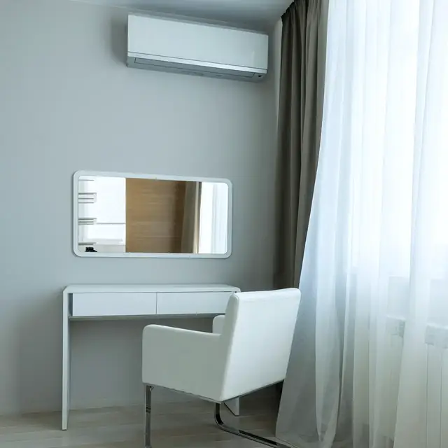 A minimal white bedroom with a dressing table, an armchair, sheer curtains and a slim wall-mounted aircon
