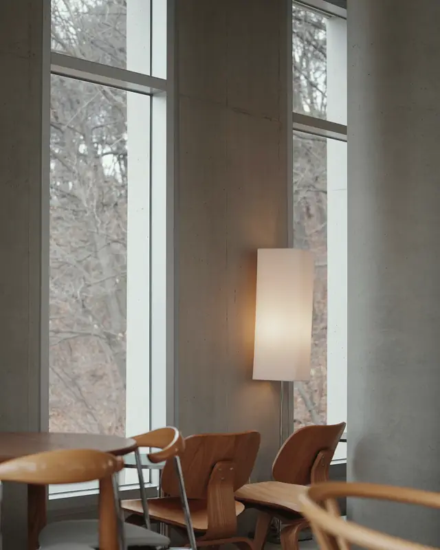 Café with tall bare windows, a concrete wall, a paper floor lamp and moulded timber chairs