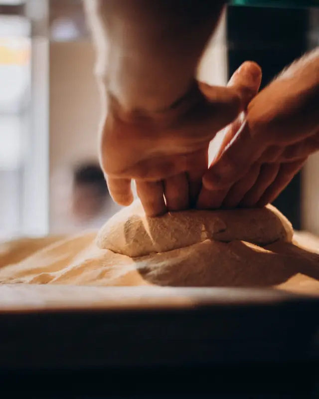 A baker's hands kneading dough on a floured wooden bench