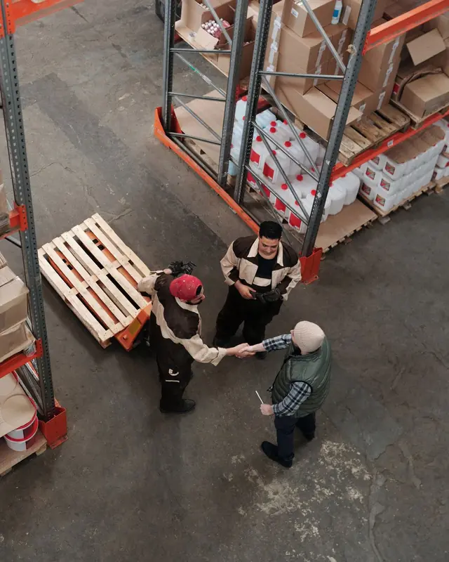 Three warehouse colleagues shaking hands beside racking and a pallet, seen from above
