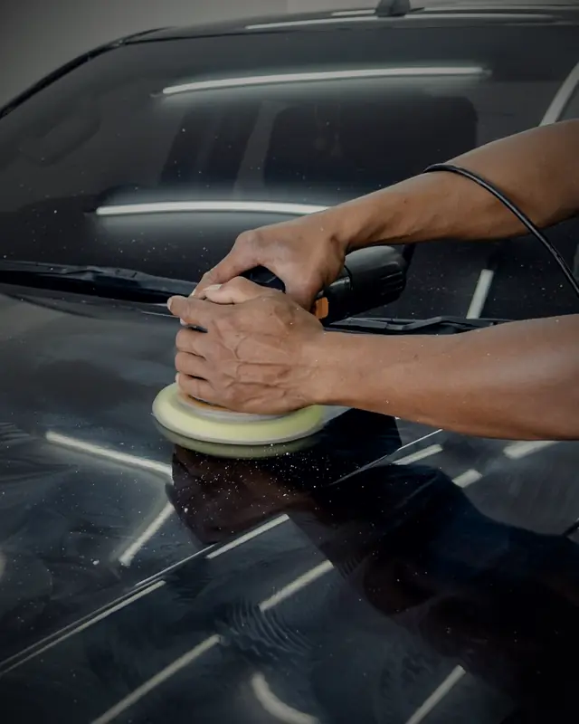 A detailer machine-polishing a black bonnet, polish residue catching the workshop light