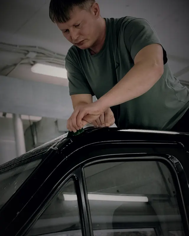 A technician fitting window film along the top of a black car's door frame