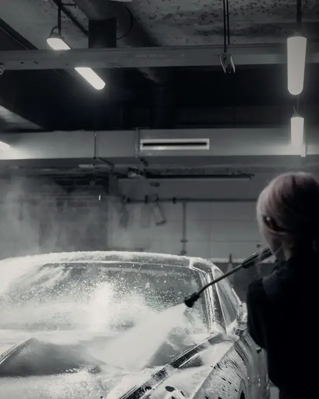 A detailer spraying snow foam over a dark car's windscreen inside the studio, under linear ceiling lights
