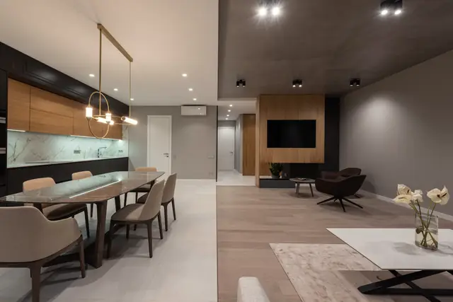 Open-plan dining and living room with a long dining table, a linear brass pendant, walnut kitchen cabinets and a timber TV wall