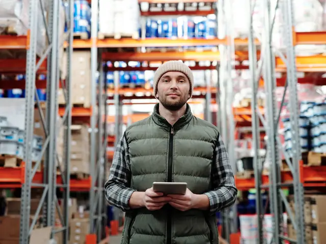 A warehouse worker in a padded vest holding a tablet between stocked shelves