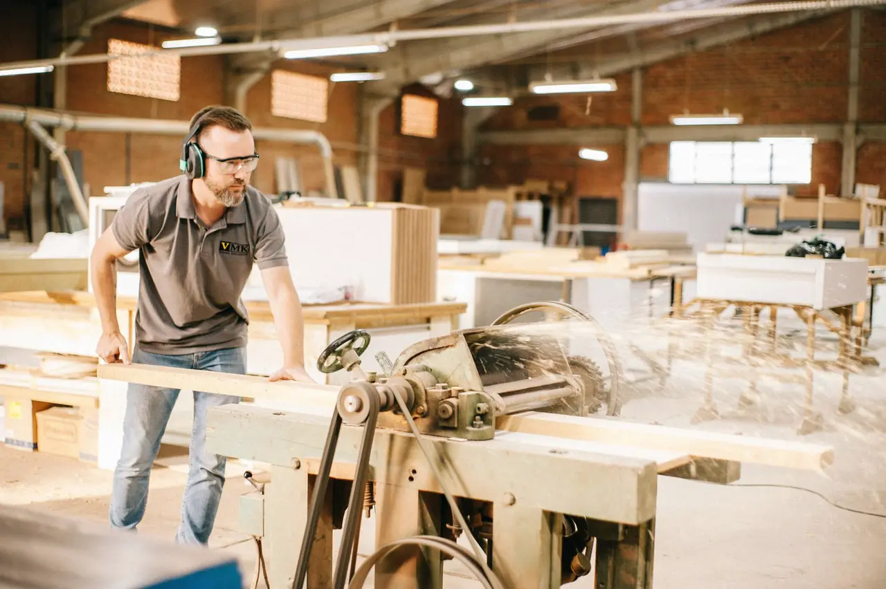 Carpenter in ear defenders working at a saw in a large workshop full of cabinet parts