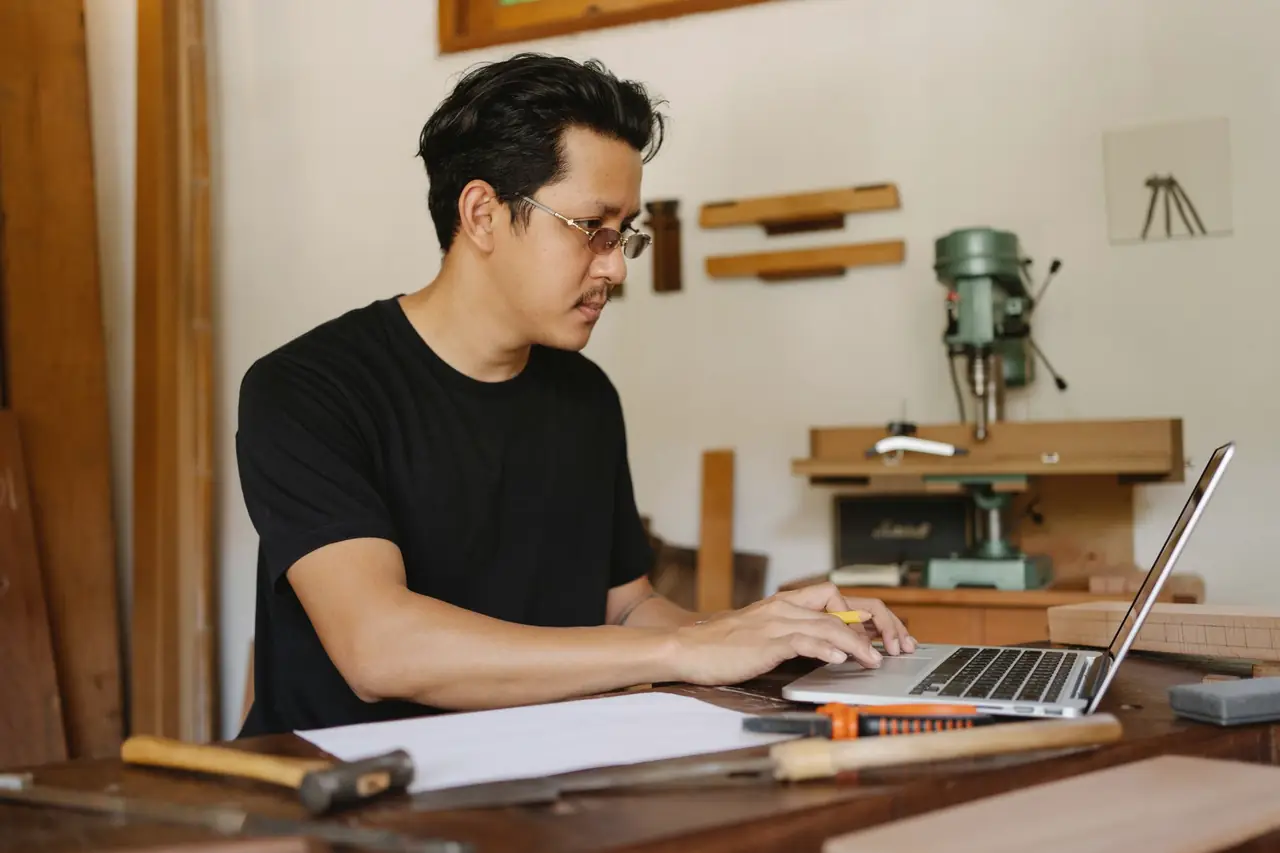 Joiner working on a laptop at a workbench, with clamps and a drill press behind him