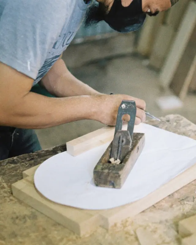 Hakim planing a pale board with a wooden hand plane at a dusty bench