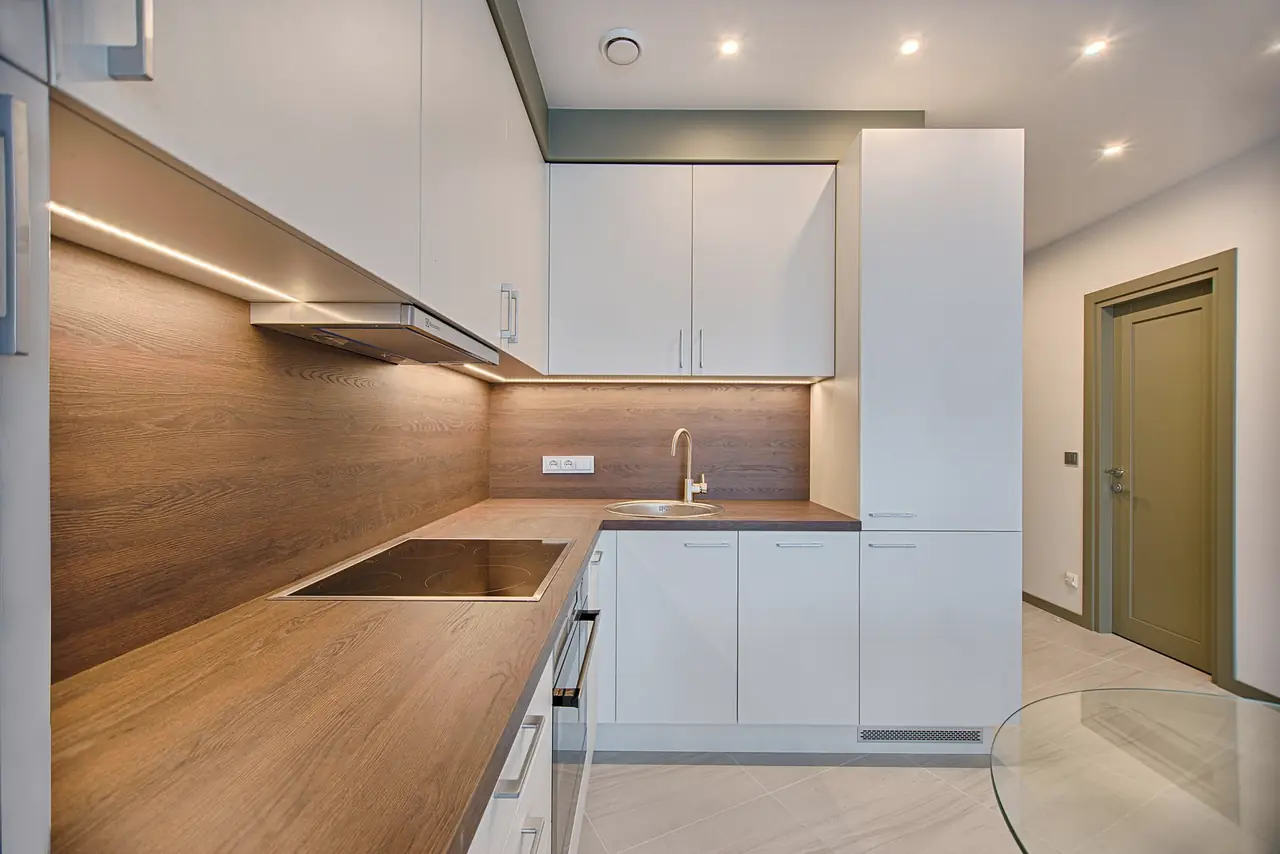 Compact L-shaped kitchen with white laminate cabinets, oak-grain worktop and backsplash, and under-cabinet lights