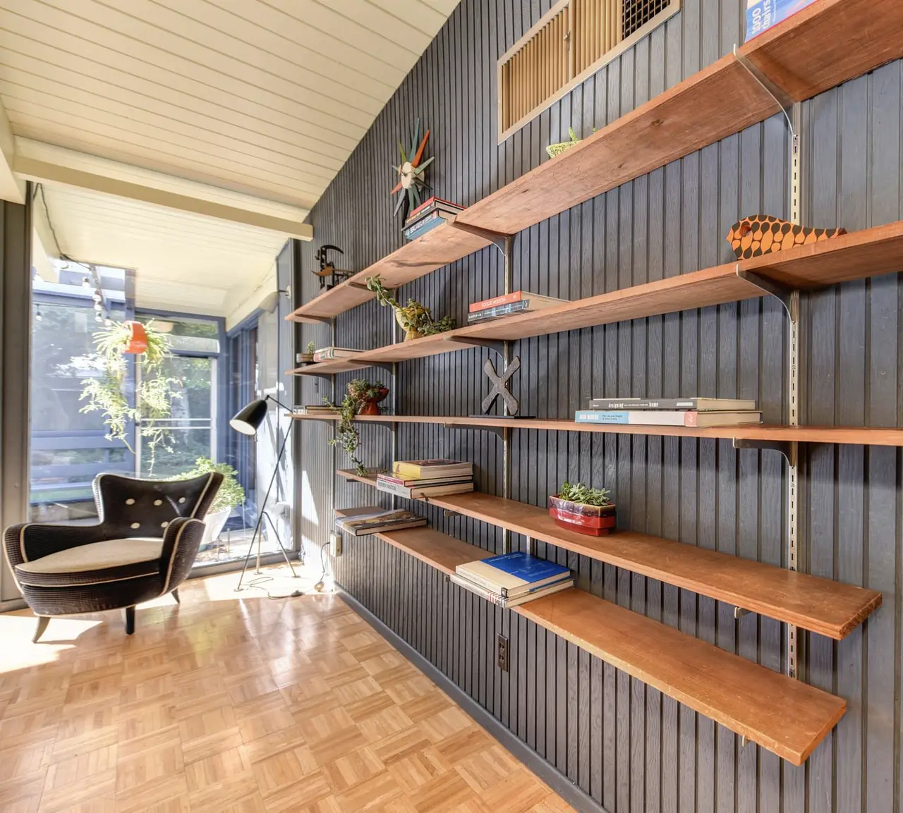 Long wall of wooden shelves on a grey slatted panel above a parquet floor, with a reading chair by the window