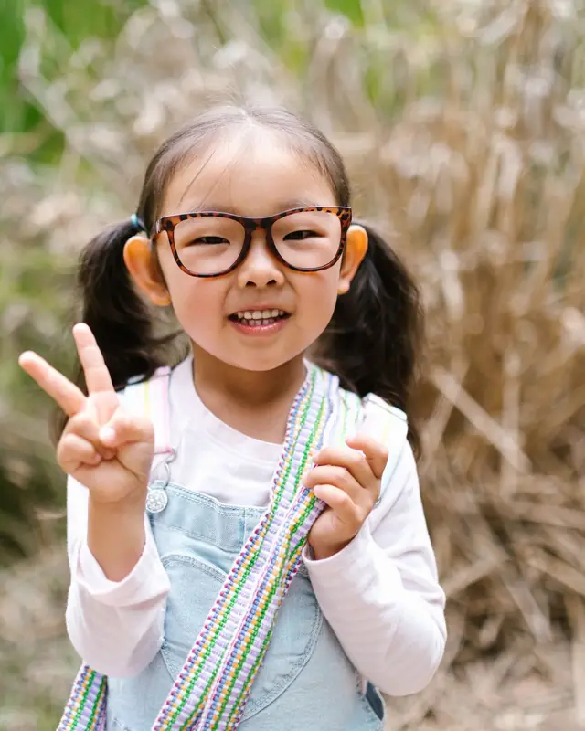 A smiling young girl in round tortoise glasses makes a peace sign outdoors