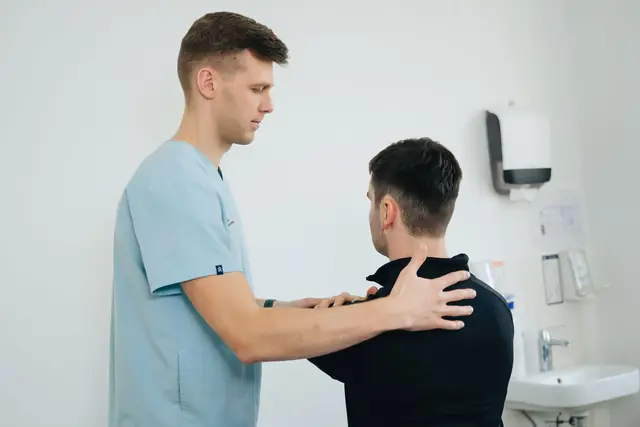 A physiotherapist stands behind a seated man with both hands on his shoulders, checking how the shoulders move