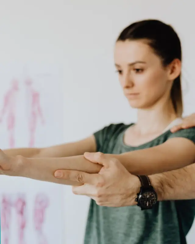 A physiotherapist steadies a woman's arms as she pulls a teal exercise band, with an anatomy chart on the wall behind