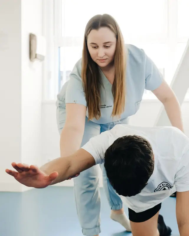 A physiotherapist guides a man through a kneeling arm-reach exercise on the floor of a bright gym room