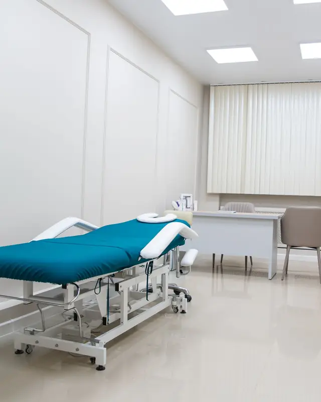 An empty treatment room with a teal treatment bed, a desk and a window with blinds