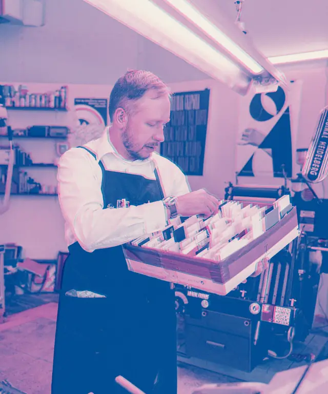A printer in an apron sorting a tray of freshly printed cards beside the press