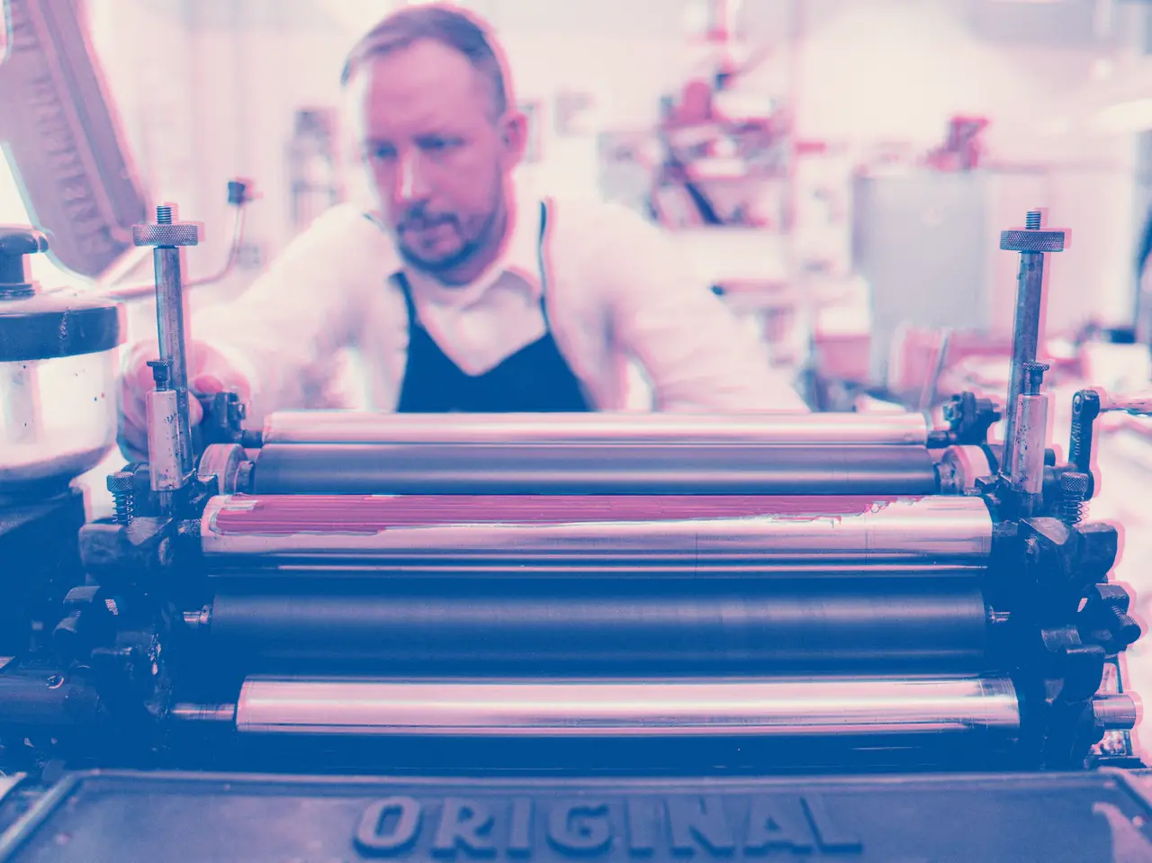Printer at work behind a vintage press, its rollers freshly inked, shown as a pink and blue two-colour print
