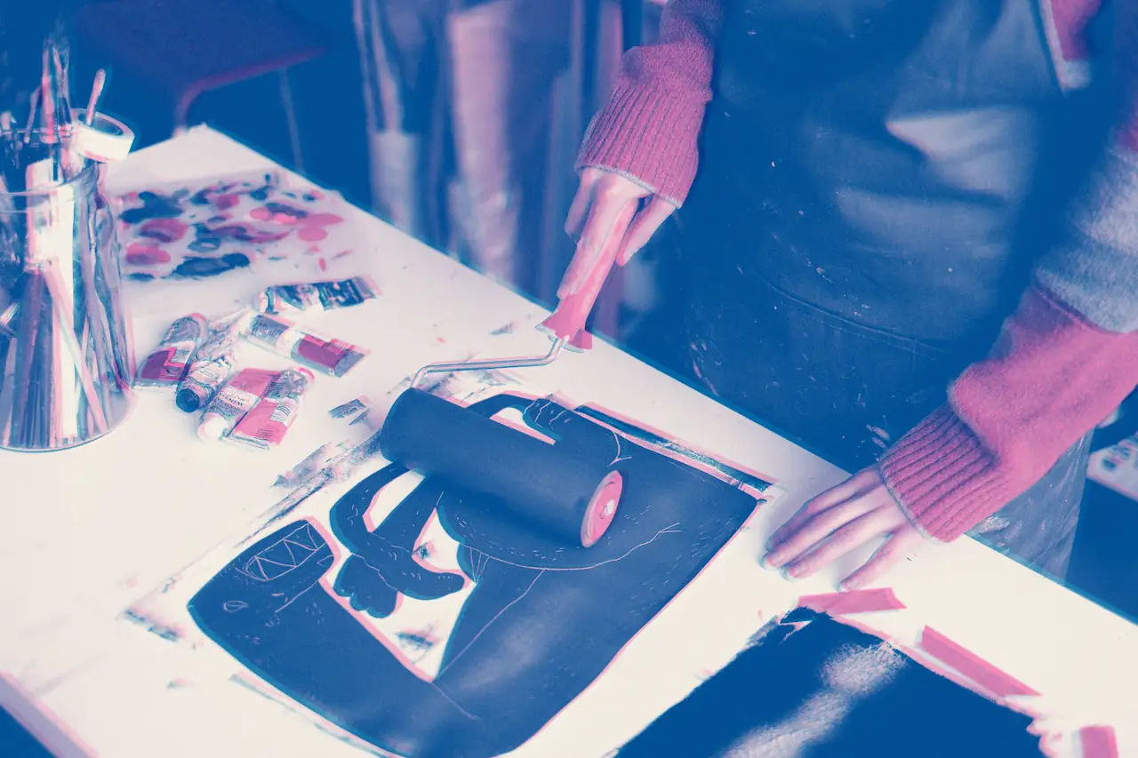 Hands rolling blue ink over a carved print block on a work table, beside tubes of ink