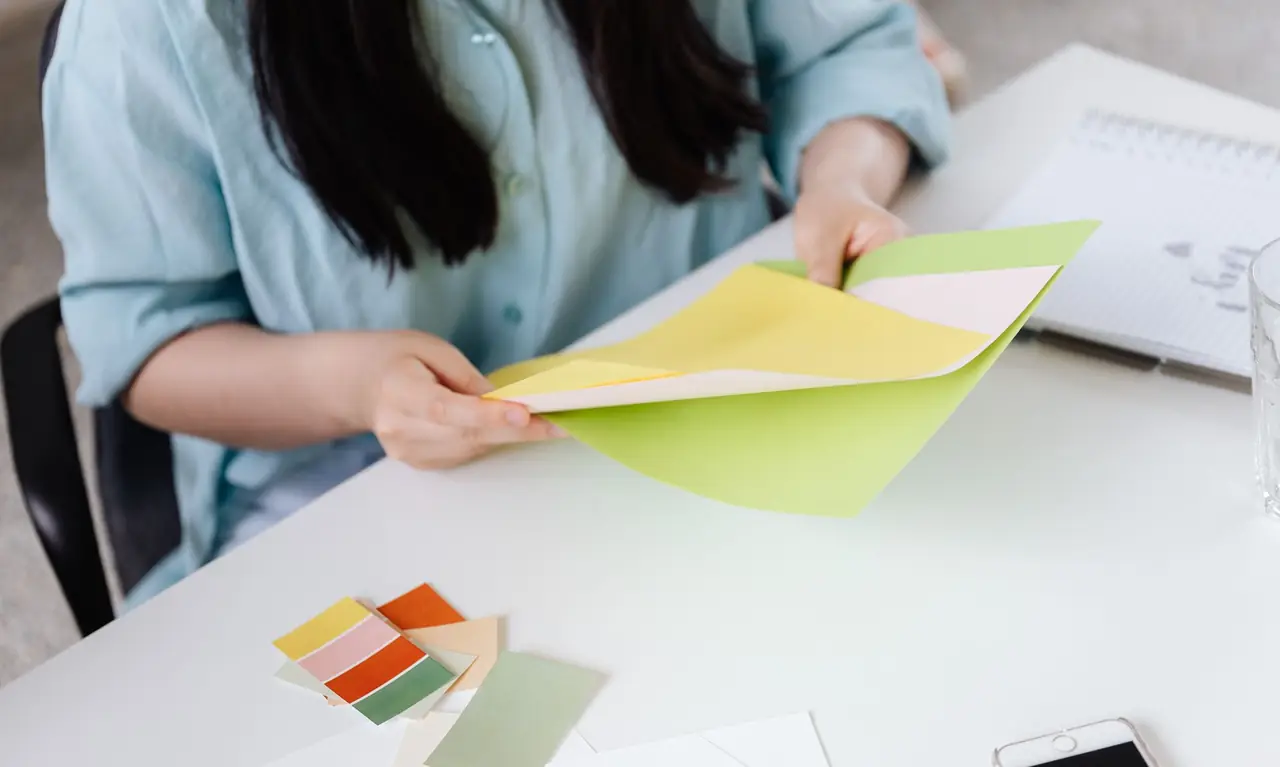 A designer at a white table holding yellow and green colour sheets, with paint chips in front of her