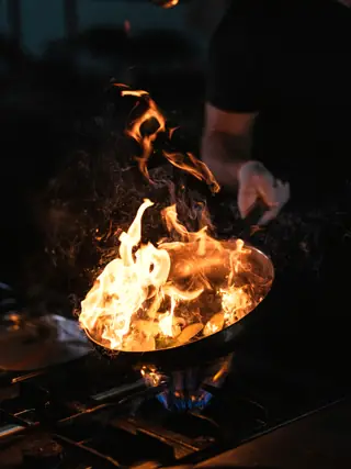 A hand shaking a flaming pan over the burner