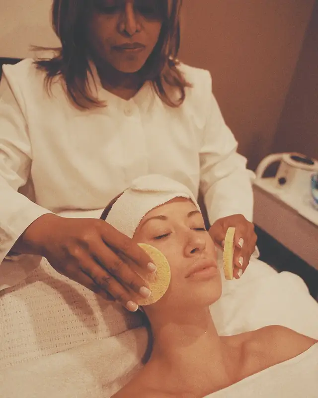 The therapist cleansing a guest's cheeks with two damp round sponges.