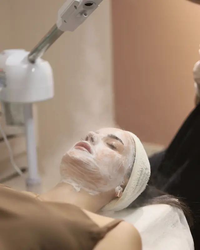 A guest lying under a facial steamer, steam drifting across her face.