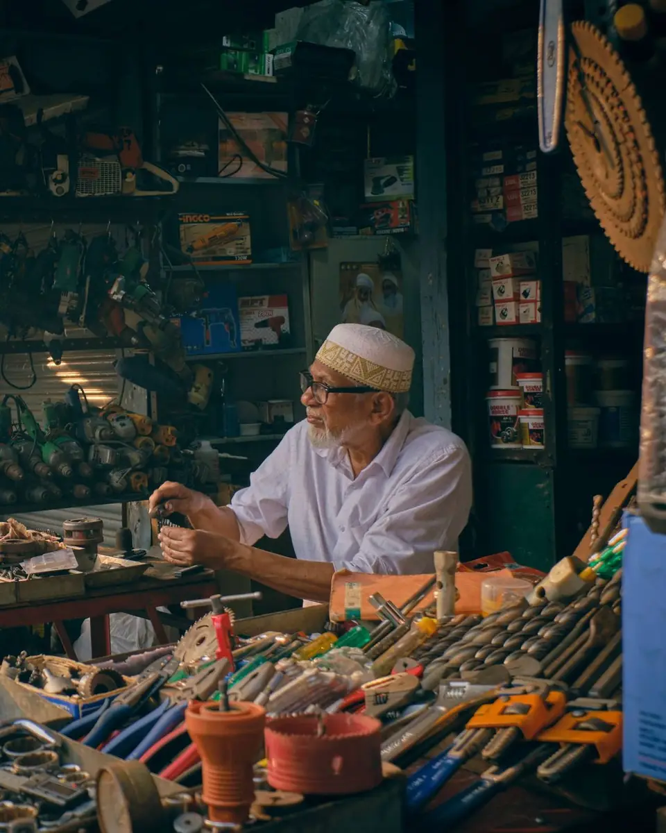 A hardware shopkeeper sorting tools at a counter crowded with fittings, hand tools and small parts