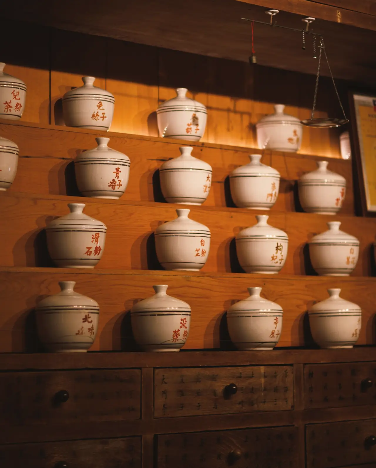 Rows of white porcelain herb jars with red hand-painted labels on warm wooden shelves above old herb drawers