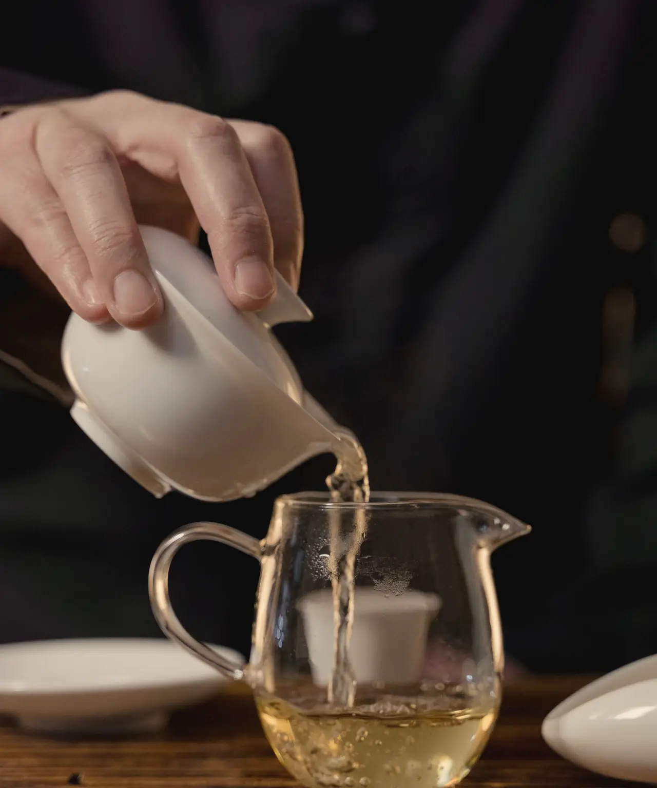 Golden tea poured from a white porcelain gaiwan into a glass pitcher on a dark wooden tea tray