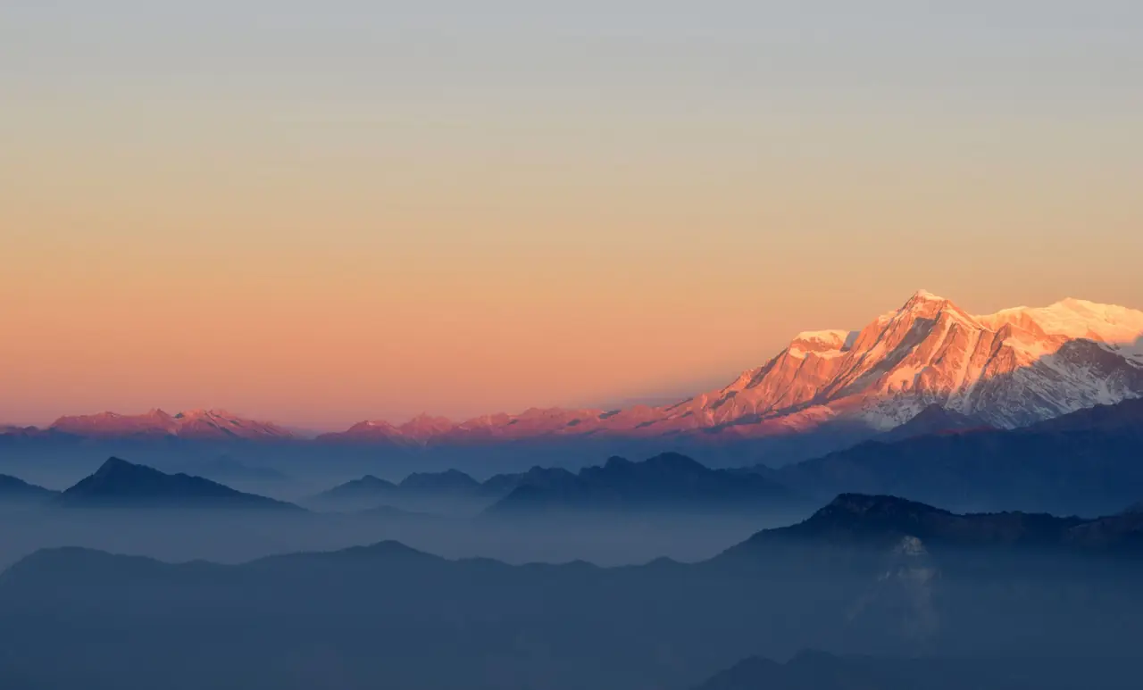 Snow peaks glowing pink and orange at sunrise above layers of blue ridges and valley mist in Nepal