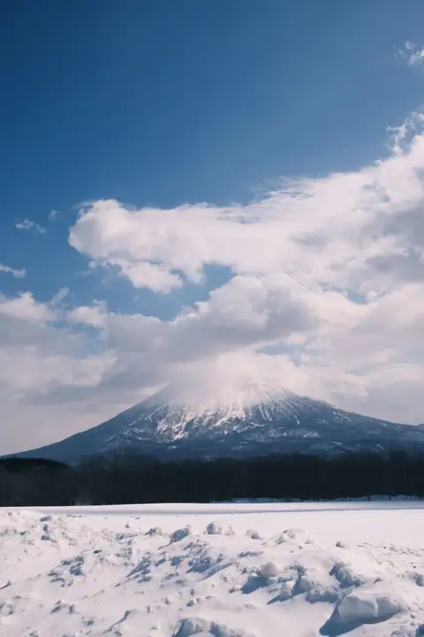 Snow-capped Mount Yotei under towering clouds and a deep blue sky, above a wind-carved snowfield in Hokkaido
