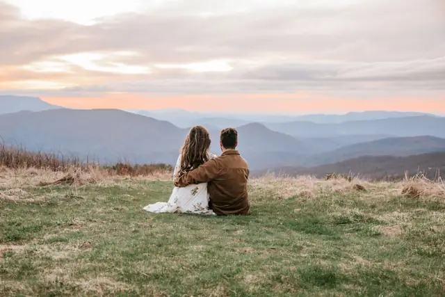 A couple sitting close together on a grassy hilltop, watching the sun set over layered mountain ridges