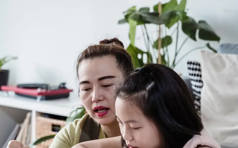 A teacher reading with a young girl at a table at home, an open book and pencil case between them