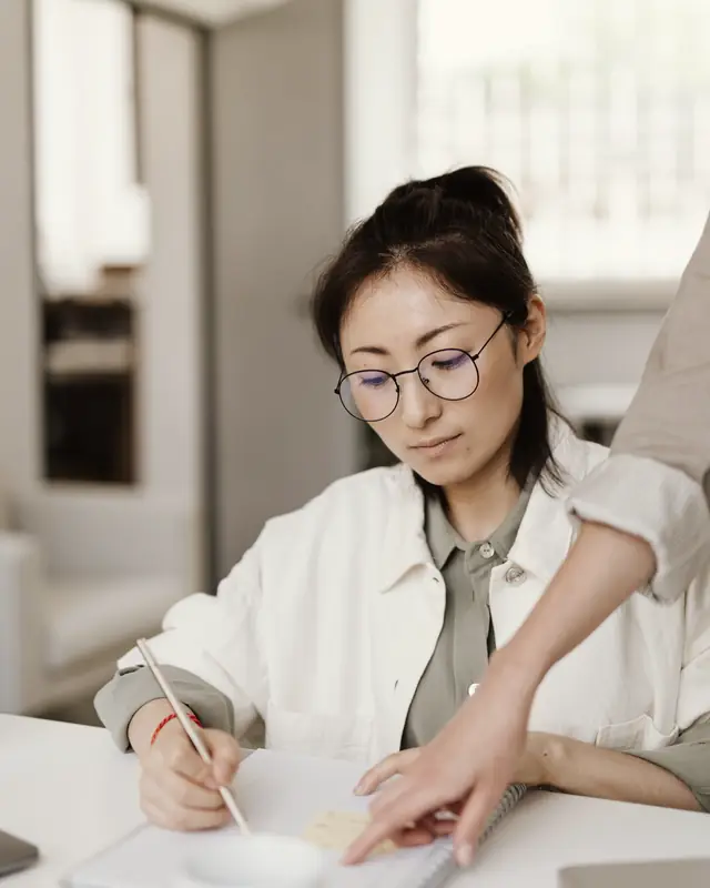 A student in glasses writing in an exercise book while her tutor points to a step in the working