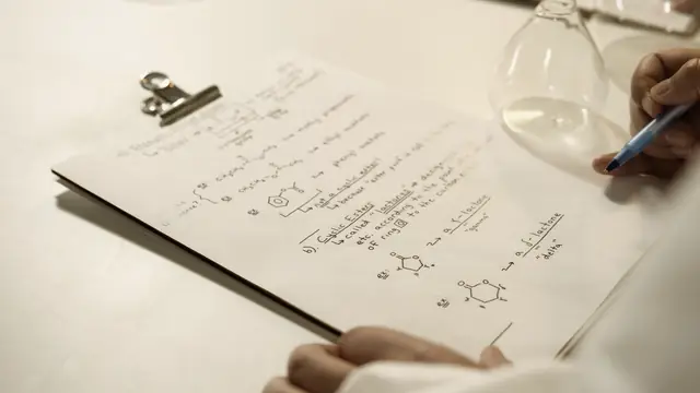 Handwritten chemistry notes with skeletal formulae on a clipboard, beside a glass flask