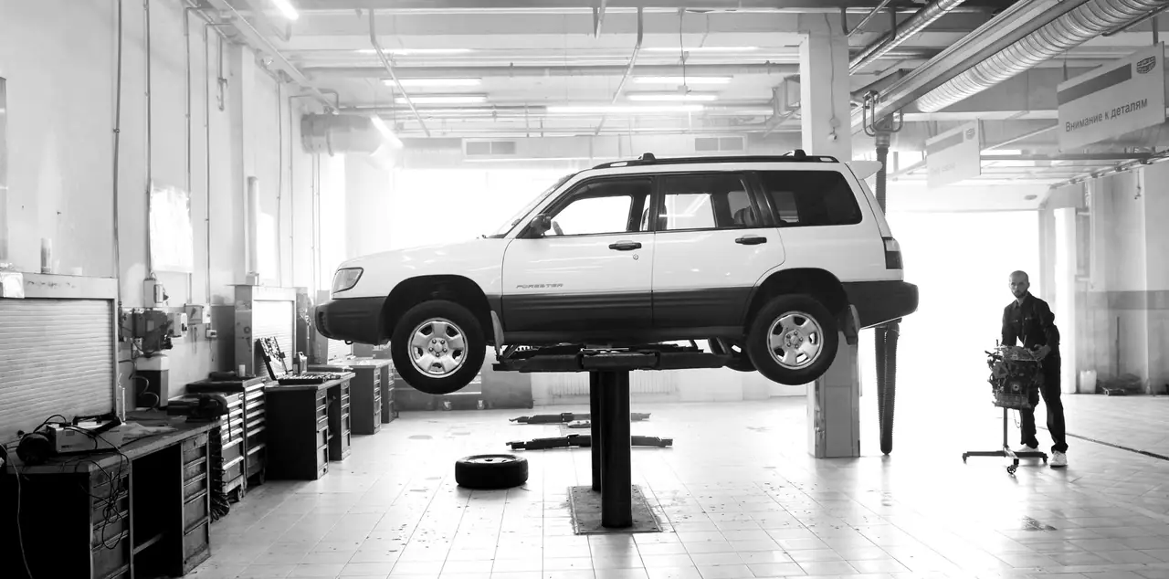 Black and white photo of the workshop floor: a white SUV raised on a two-post lift, tool chests along the wall, a technician walking past
