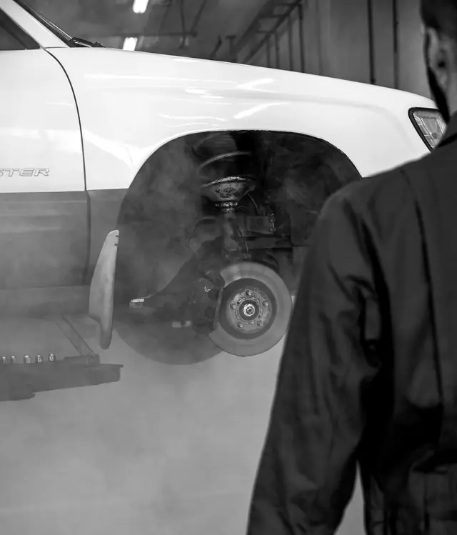 A technician walks towards a raised SUV with its front wheel off, the brake disc and spring exposed